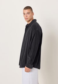 Mladić s kratkom kosom, odjeven u labavu crnu košulju od manšestera i svijetlosive hlače, stoji nasuprot običnoj svijetloj pozadini.