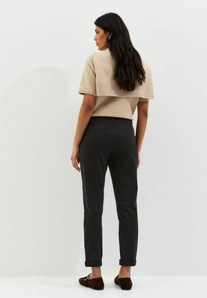 Mujer con cabello largo y oscuro, vestida con una blusa beige de manga corta y pantalones negros remangados, posando frente a un fondo blanco liso.