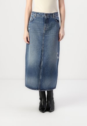 Vrouw draagt een vervaagde blauwe denim midirok met lichte slijtage en zwarte laarzen met puntige neus en hakken op een witte achtergrond.