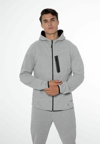 Haut zippé gris avec des accents noirs, doté d'une poche avant zippée et d'un tissu texturé. Le modèle se tient les bras croisés, portant un pantalon de jogging assorti.