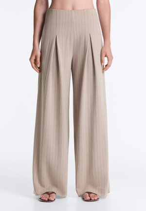 Pantalon beige à jambes larges avec des rayures verticales et des plis devant, porté avec des sandales marron, présenté de la taille aux pieds sur un fond uni.
