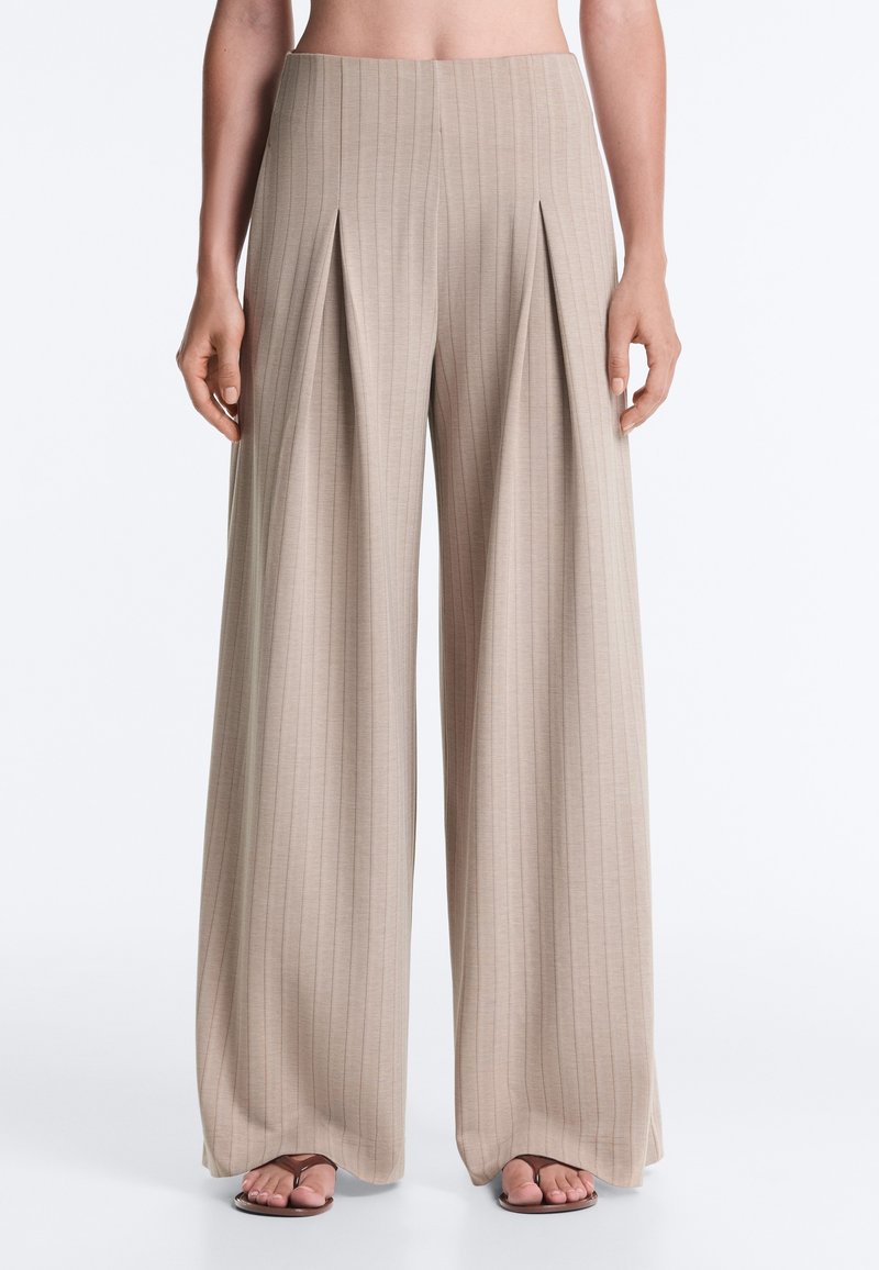Pantalon beige à jambes larges avec des rayures verticales et des plis devant, porté avec des sandales marron, présenté de la taille aux pieds sur un fond uni.