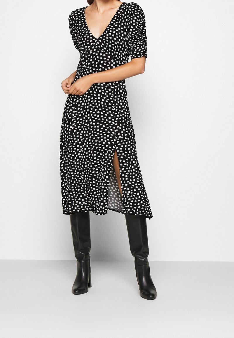 Žena v černých midi šatech s bílými puntíky a bočním rozparkem, v kombinaci s černými vysokými kozačkami na podpatku, stojící na bílém pozadí.