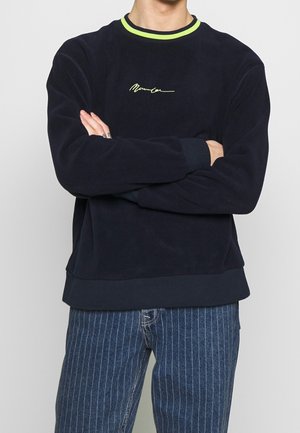 Mand iført marineblå sweatshirt med neongult kantbånd og kursivlogo, kombineret med blå jeans med smalle striber, armene krydsede.