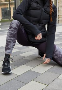 Persona estirando la pierna en una acera pavimentada, vestida con ropa deportiva negra y morada y zapatos de correr negros.