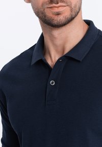 Marinblå polo tröja, kortärmad, tillverkad av texturerad tyg. Har en krage och en knappslå med detaljer i svart knappland.