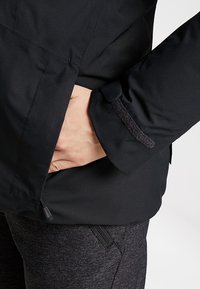 Svart vattentålig jacka med en texturerad yta, med en dragkedjeförsedd handficka och justerbara ärmar. Kombinerad med mörkgrå byxor.