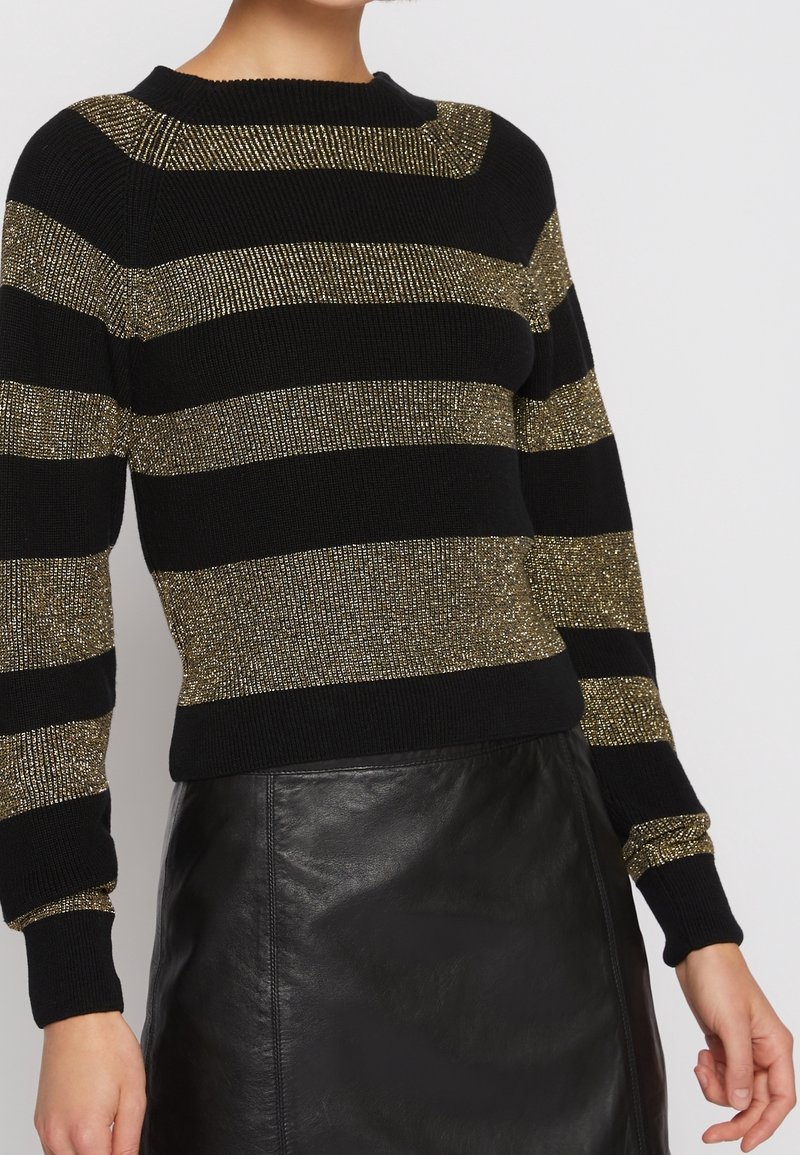 Femme portant un pull tricoté rayé noir et or métallisé associé à une jupe en cuir noire sur un fond uni.