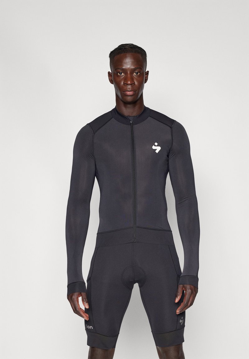 Body de sport noir avec manches longues, fermeture éclair sur le devant et shorts ajustés. Logo notable sur la poitrine et la jambe, texture lisse tout au long.