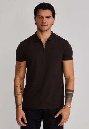 Man met kort donker haar, gekleed in een donkerbruin poloshirt met korte mouwen en een gouden rits, met zichtbare tatoeages op beide armen.