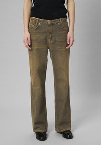 Brązowe dżinsy o luźnym kroju, pięciokieszeniowym wzorze i subtelnych zagięciach. Posiadają wysoki stan i prosty krój nogawki, zestawione z czarnymi butami.