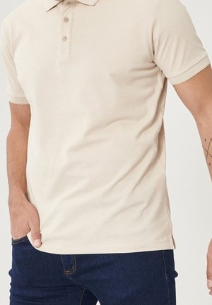 Mand iført beige kortærmet polo-shirt og mørkeblå jeans, med den ene hånd i lommen og tatovering på højre underarm.