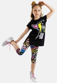 Schwarzes T-Shirt mit buntem Blitzgrafik, kombiniert mit Capri Leggings in lebhaftem Tierdruck. Silberne Sneakers mit pinken Schnürsenkeln.