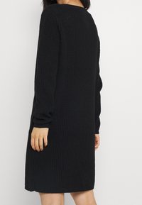 Svart stickad klänning med långa ärmar, ribbad textur och avslappnad passform. Fållen är något ovanför knäet och har en enkel design.