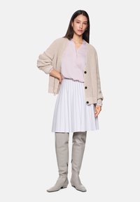 Beige gestrickte Cardigan, hellrosa Bluse, weiße plissierte Bluse und hohe graue Stiefel. Das Outfit hat Knöpfe und strukturierte Materialien.