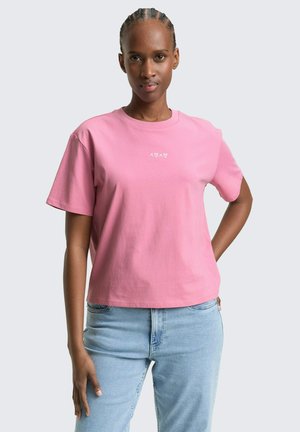 Kobieta z zaplecionymi włosami, ubrana w różową koszulkę z małymi białymi wzorami serc i liter X oraz jasnoniebieskie dżinsy, stojąca z jedną ręką na biodrze.