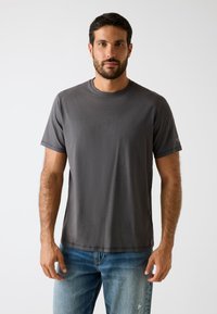 Graues kurzärmeliges T-Shirt aus weichem Stoff, mit Rundhalsausschnitt und einem dezenten Logo auf dem linken Ärmel. Kombiniert mit hellblauen Jeans.