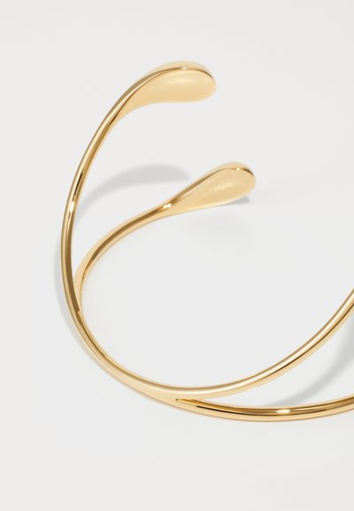 Pulsera de brazalete de metal dorado con un diseño liso y curvado, con extremos puntiagudos; superficie brillante; estética minimalista y moderna.