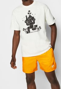 Biała koszulka z czarnym graficznym wzorem przedstawiającym abstrakcyjne kształty i roślinność. W połączeniu z pomarańczowymi szortami z logo Nike.