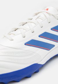 Zapatilla deportiva blanca con parte superior sintética texturizada, acentos de goma azul, tres rayas azules y detalles rojos en la parte superior.