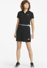 Vestido polo negro con mangas cortas, logo blanco de Puma en el pecho y en la falda, y una cinturilla con un patrón en blanco y negro. Combinado con zapatos deportivos negros.