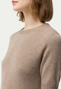 Brun stickad tröja med rund halsringning och korta ärmar, som visar en mjuk textur och en figurnära siluett. Sömdetaljer är synliga.