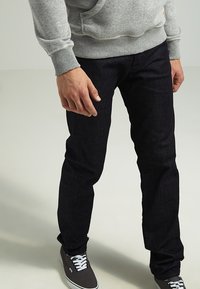 Jeans in denim blu scuro con una vestibilità slim, abbinati a un maglione grigio. I jeans presentano una leggera trama e sono risvoltati alle caviglie.
