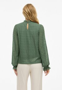 Groene kant blouse met lange, opgeblazen mouwen en een gerimpelde halslijn. Heeft een sleutelgatopening aan de achterkant en textuurpatronen van bloemen.