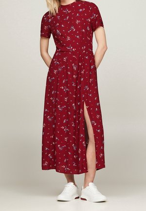 Femme portant une robe midi rouge à fleurs avec des manches courtes et une fente latérale, associée à des baskets blanches, debout les mains dans le dos.