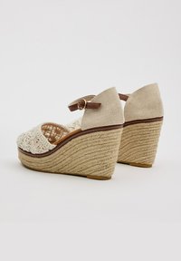 Sandalias de cuña con parte superior de tejido blanco, tira marrón con hebilla y base de cuerda texturizada. Presentan un diseño de talón cerrado y punta abierta.