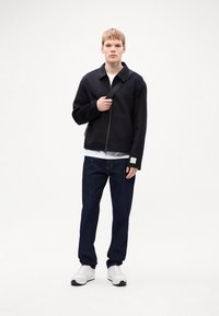 Veste noire zippée avec un col, associée à un t-shirt blanc et un jean bleu foncé. Des baskets blanches complètent ce look décontracté.