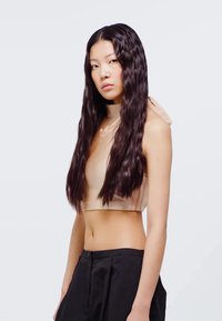 Beige cropped linne med hög halsringning och öppen rygg. Modellen bär svarta, löst sittande byxor. Långt, vågigt hår faller över axlarna.