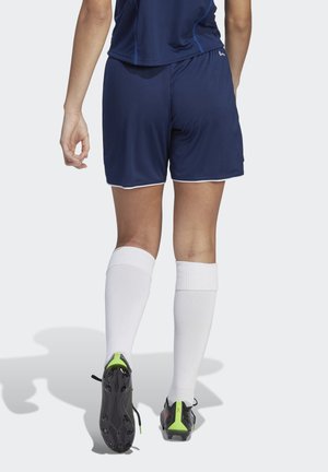 Marineblauwe shorts met witte bies, gecombineerd met kniehoogte witte sokken. Zwarte en groene schoenen met studs op de zool.