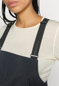 Σκούρες γκρίζες σαλοπέτες με ρυθμιζόμενες τιράντες που φέρουν λογότυπο. Φοριέται πάνω από μια ανοιχτού κρεμ μπλούζα με στρογγυλή λαιμόκοψη, που εμφανίζει ομαλή υφή.