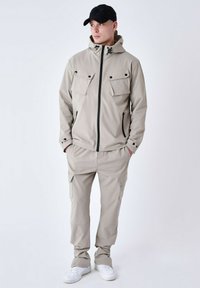 Chaqueta ligera beige con dos bolsillos en el pecho, cremallera negra y capucha ajustable, combinada con pantalones cargo a juego y zapatillas blancas.