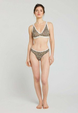 Femme debout pieds nus portant un soutien-gorge à imprimé léopard et une culotte taille haute assortie sur un fond clair uni.