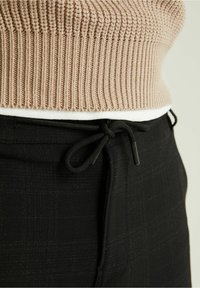 Beige pletený svetr zasunutý do černých kalhot s viditelným černým šňůrkovým páskem na pasu na čistém pozadí.