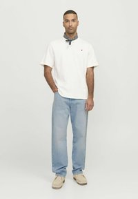 Camisa blanca de manga corta con logo rojo, jeans rectos de color azul claro y zapatos beige. Complementa con un pañuelo oscuro de estampado alrededor del cuello.