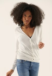 White cardigan with a V-neckline and button closure, made of soft material. Paired with blue jeans, featuring a slim fit and medium wash.