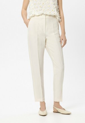 Frau trägt hoch taillierte, cremefarbene maßgeschneiderte Hose mit vorderen Falten, kombiniert mit cremefarbenen Slingback-Ballerinas und einem weißen, mit Margeriten verzierten Oberteil.