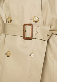 Beige trenchcoat med matchande tygskärp med brun rektangulär spänne och fyra beige knappar med texten "Lauren Ralph".