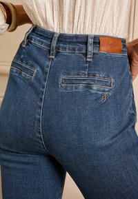 Temno modri džins hlače s tesno prilegajočo zasnovo, ki vključuje usnjeno oznako na pasu in dve zadnji žepni z vodoravnimi odprtinami.