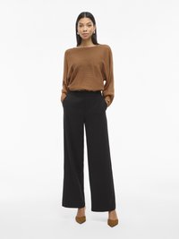 Femme portant un pull côtelé marron, un pantalon large noir et des talons aiguilles marron, se tenant les mains dans les poches sur fond blanc.
