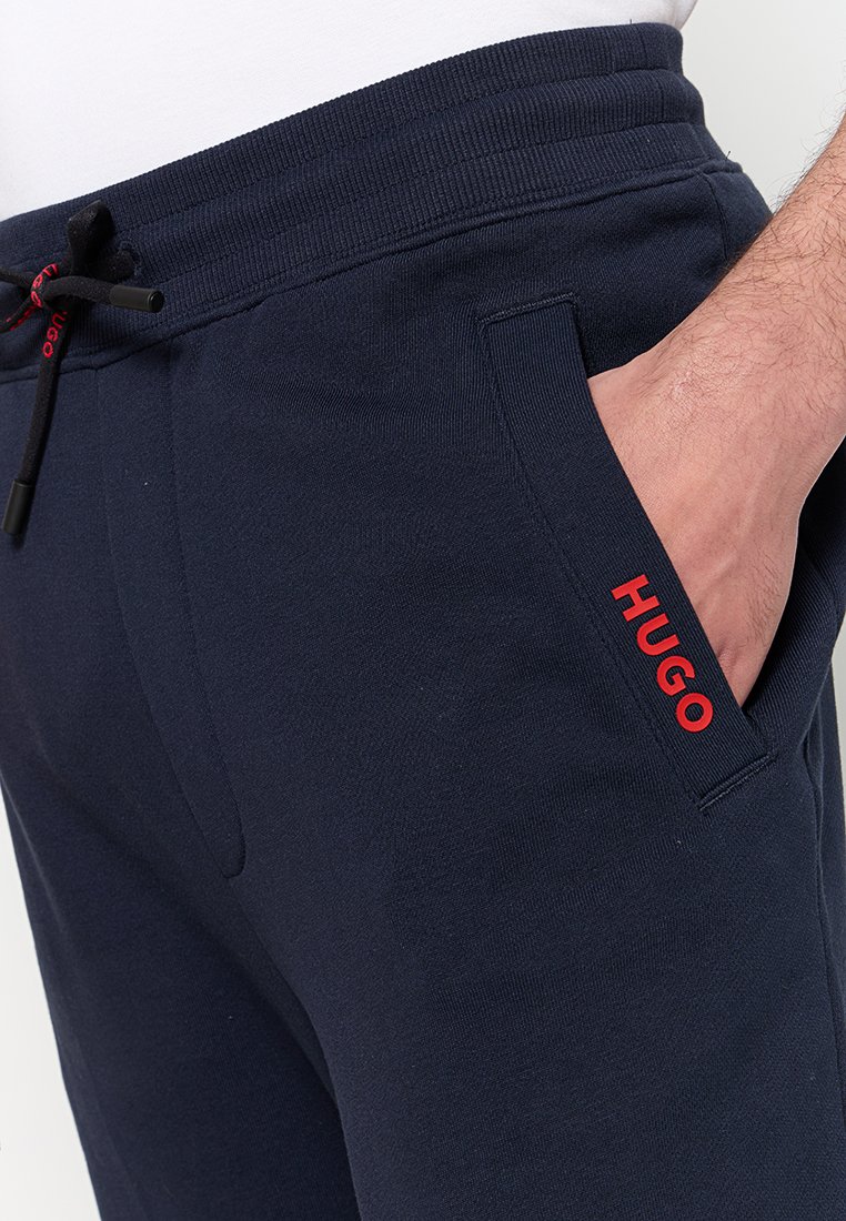 Mężczyzna w granatowych spodniach dresowych z elastycznym paskiem i czarnym sznurkiem, ręka w kieszeni, czerwone logo "HUGO" na krawędzi kieszeni.
