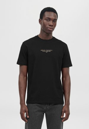 Мъжки модел стои на светъл фон, облечен с черна тениска Armani Exchange и тъмносиви дънки.