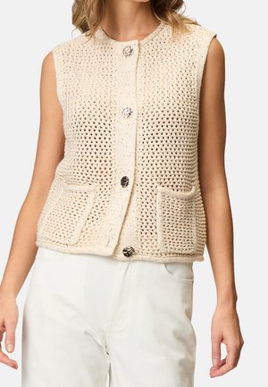 Veste beige sans manches en crochet avec de gros boutons et des poches avant portée sur un pantalon blanc.