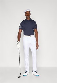 Marineblå poloshirt med mønstrede ærmer, hvide bukser, hvide golfsko med blå detaljer og en hvid cap. Holder en golfkølle.