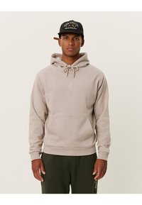 Felpa con cappuccio beige chiaro, dotata di coulisse, tasca frontale e branding minimal. Tessuto morbido e vestibilità regolare, indossata con pantaloni verde scuro.