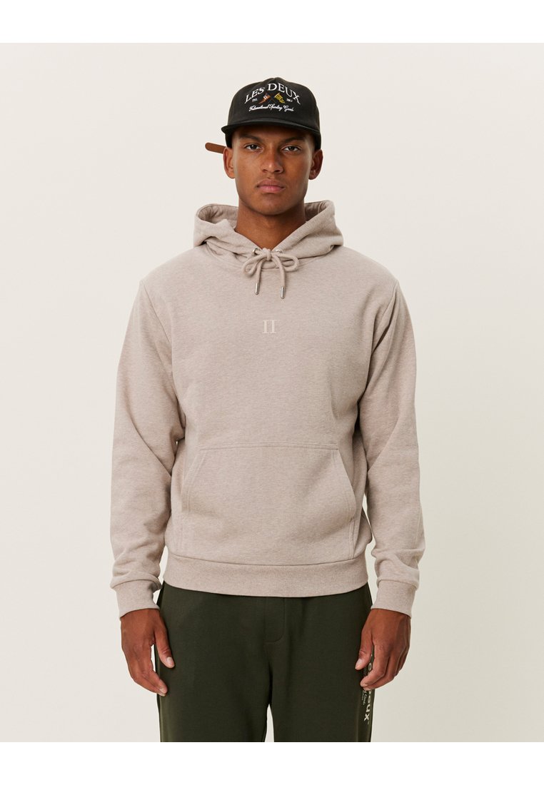 Felpa con cappuccio beige chiaro, dotata di coulisse, tasca frontale e branding minimal. Tessuto morbido e vestibilità regolare, indossata con pantaloni verde scuro.