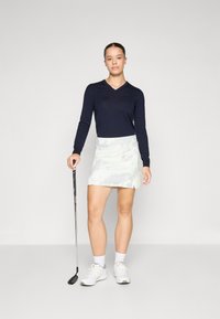 Marinblå långärmad v-ringad tröja kombinerat med en ljusmönstrad golfkjol. Vita sportskor och en golfklubba i handen.